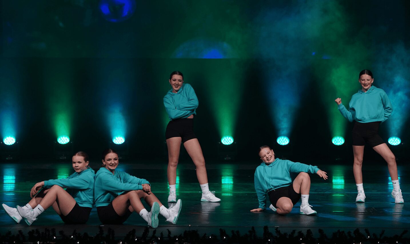 Five teal-hooded dancers posing on lit stage